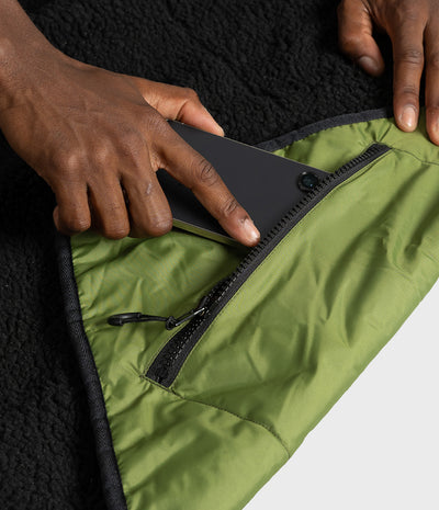 Close-up of a person's hands demonstrating a zippered pocket on an olive green picnic blanket, with a black fleece lining visible underneath.