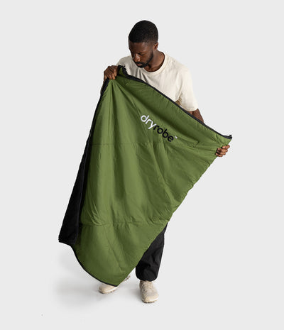 A man wearing a beige t-shirt and black pants holds up a green picnic blanket with a black interior lining and the 'dryrobe' logo printed on it, against a light gray studio background.