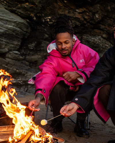 *MALE* A person wearing a bright pink hooded jacket sits beside a campfire outdoors, roasting a marshmallow on a stick. Another person in a dark jacket is partially visible at the right edge, with rocky terrain in the background.