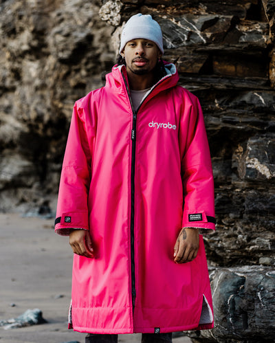 *MALE* A man wearing a bright pink dryrobe hooded raincoat and a white beanie stands in front of a dark rocky cliff background.