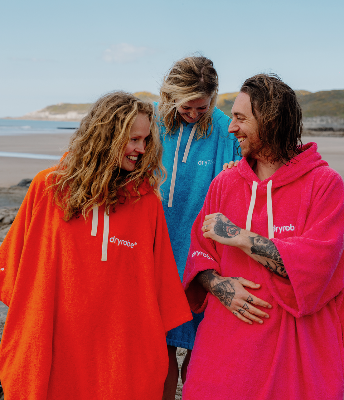 Three people wearing colorful dryrobe ponchos on a beach.
