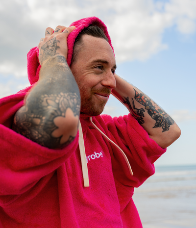 *MALE* Man wearing a pink hooded towel robe with 'dryrobe' branding, standing outdoors against a cloudy sky.
