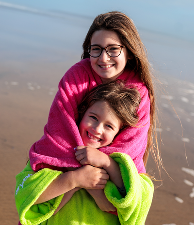 Two children hugging on a beach, one wrapped in a pink towel robe and the other in a green towel robe