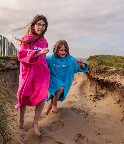 Two children in pink and blue dryrobes running on a sandy beach.