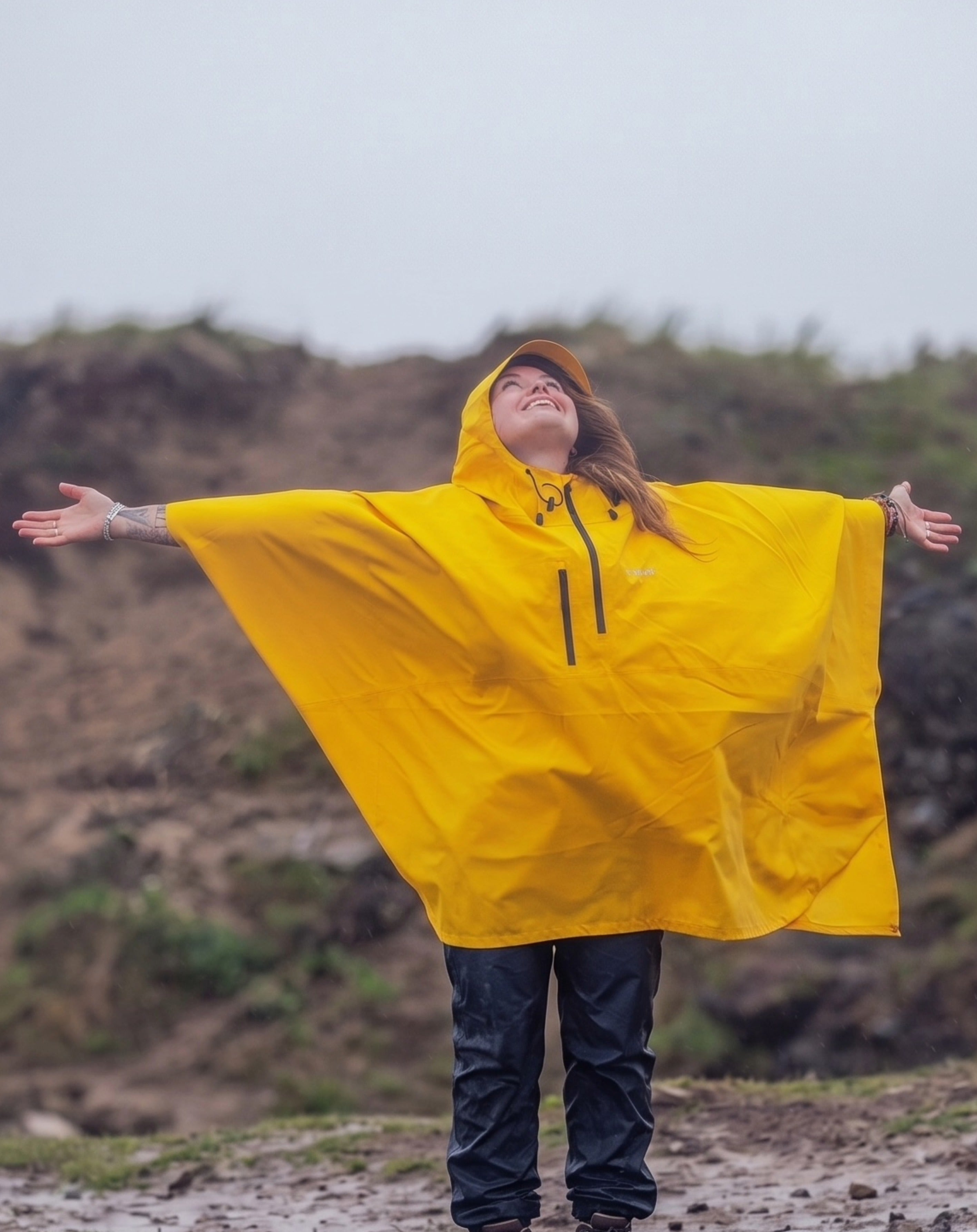 A woman wearing a bright yellow hooded rain poncho with her arms outstretched, standing outdoors on a muddy path with a rocky hillside in the background.