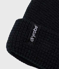 Close-up of a black waffle-knit beanie with a folded cuff, featuring a black fabric label with the brand name 'dryrobe' in white text, set against a light gray background.