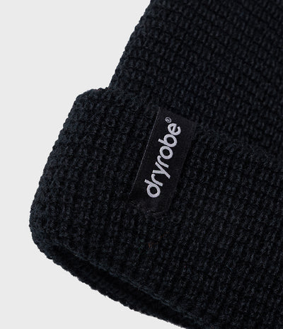 Close-up of a black waffle-knit beanie with a folded cuff, featuring a black fabric label with the brand name 'dryrobe' in white text, set against a light gray background.