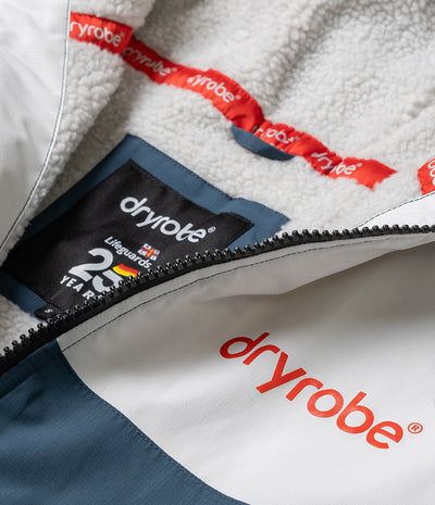 *MALE* Close-up of a Dryrobe jacket showing the white sherpa lining, a black brand label with '25 Year' text, and the white and navy outer fabric with red Dryrobe logo and zipper.