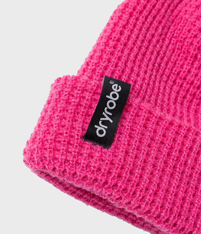 A close-up of a bright pink ribbed knit beanie with a black fabric brand tag reading 'dryrobe' in white text, set against a light gray background.