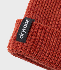 Close-up of a rust-orange ribbed knit beanie with a black fabric Dryrobe brand tag on the cuff, set against a light gray background.