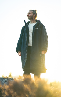 Man wearing a long dark coat standing outdoors with sunlight filtering through