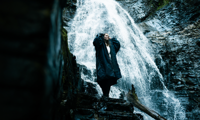 Person standing in front of a waterfall wearing a dark coat.