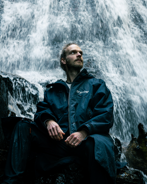Man sitting in front of a waterfall wearing a dark jacket with 'Tyr' branding.