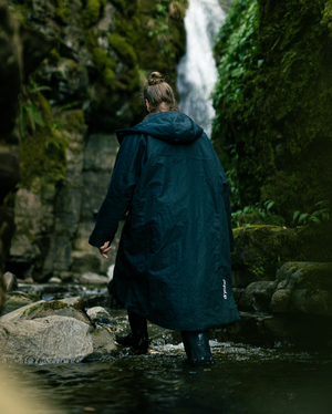 Person in a long dark coat walking towards a waterfall in a forested area