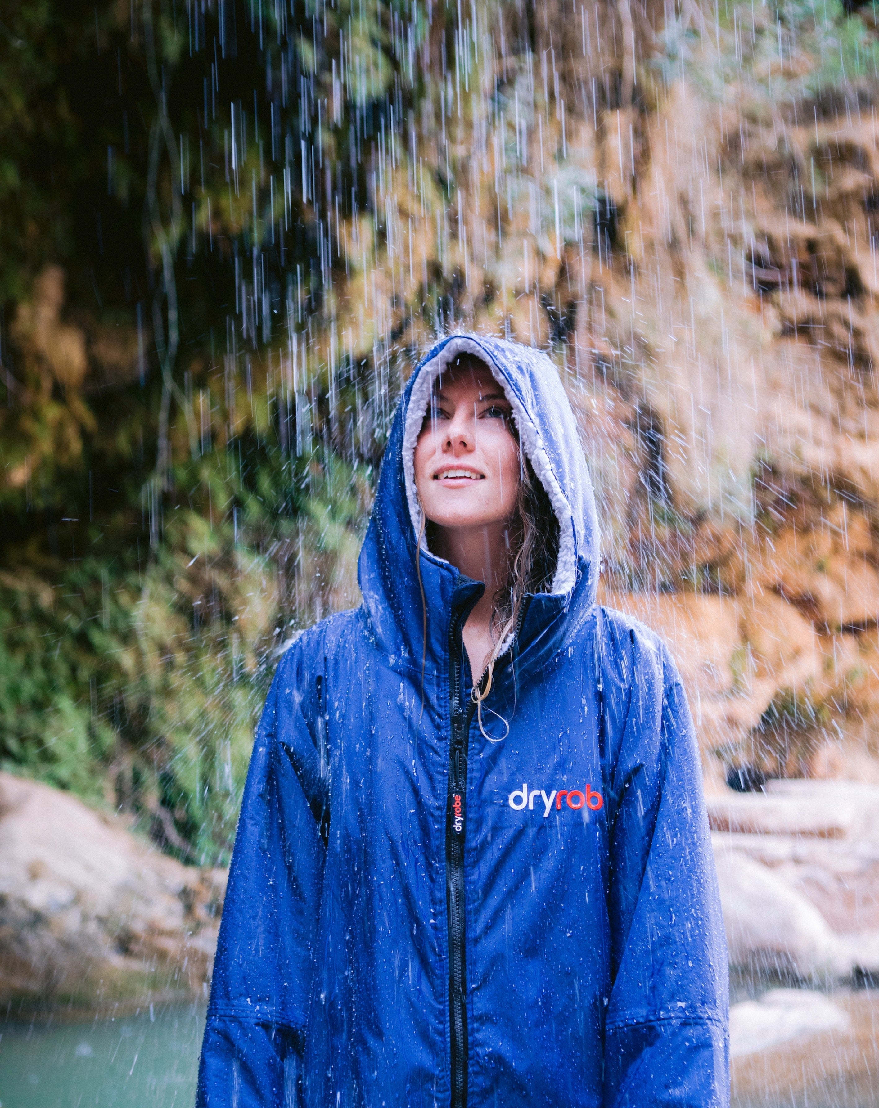 A woman wearing a blue Dryrobe stands under falling rain in an outdoor natural setting with a rocky, mossy background.