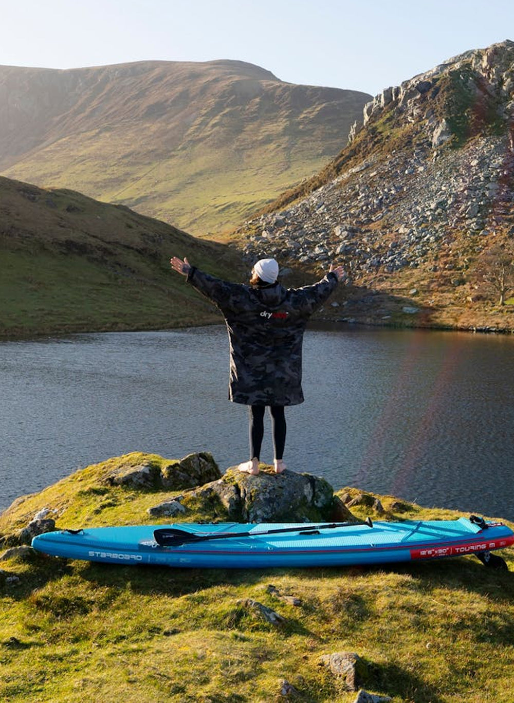 Man wearing Black Camo dryrobe advance standing with his paddleboard at the edge of a lake