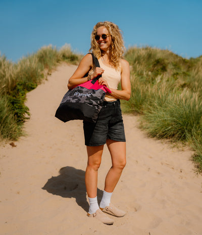 A woman stands on a sandy path between dune grass under a blue sky, carrying a black tote bag with a pink interior and the brand name 'horse' printed on it.
