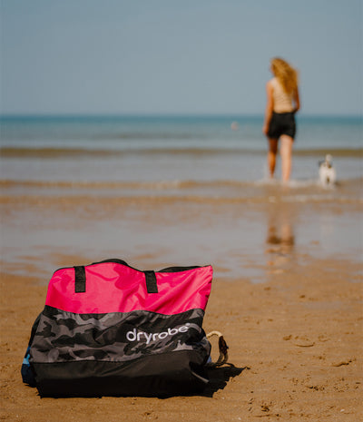 A pink and black Dryrobe dog bag sits on a sandy beach in the foreground, with a woman and her dog standing in the ocean water in the blurred background.