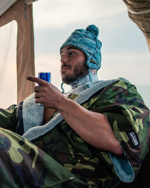 Person wrapped in a camouflage dryrobe, wearing a blue knit hat, sitting inside a tent.