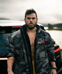 Man wearing a camouflage Dryrobe in front of a car.
