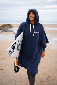 A person standing on a sandy beach wearing a navy blue hooded towel poncho with white drawstrings and carrying a white surfboard.