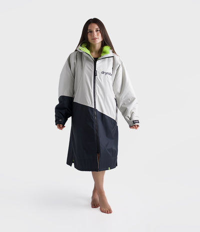 A woman modeling a colourblock hooded Dryrobe with a light grey upper section, dark navy lower section, and a bright lime green inner hood lining, standing against a light grey studio background.