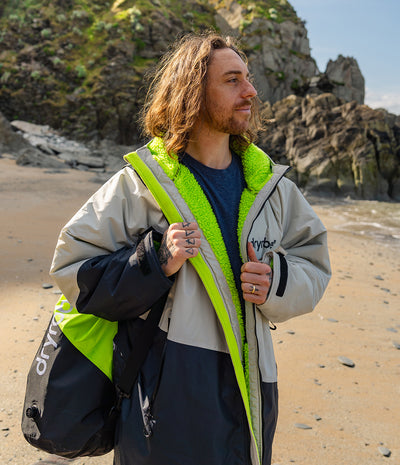 *MALE* Man standing on a beach wearing a Dryrobe with a visible brand logo, holding a bag.
