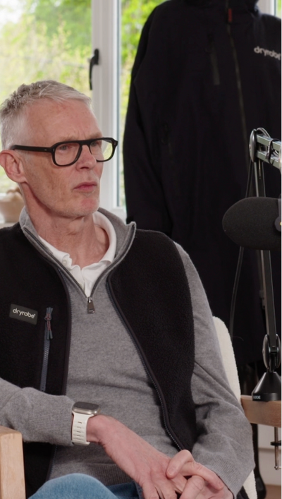 Man wearing a black jacket with a visible brand logo, sitting indoors.