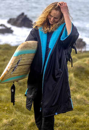 Woman smiling carrying a surfboard by the sea, wearing dryrobe® Advance Short Sleeve