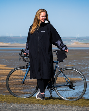 Woman in a black coat standing next to a bicycle by a coastal area with wind turbines in the background.