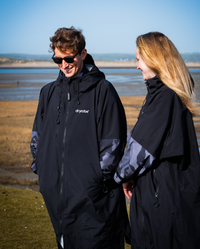 Two people wearing black jackets with camouflage patterns, standing by a coastal area.