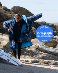 Person wrapped in a blue and navy patterned dryrobe with a surfboard, standing on rocks near the ocean.