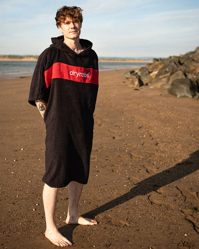 Man stood on a beach wearing Organic Towel dryrobe Remix in Black Red