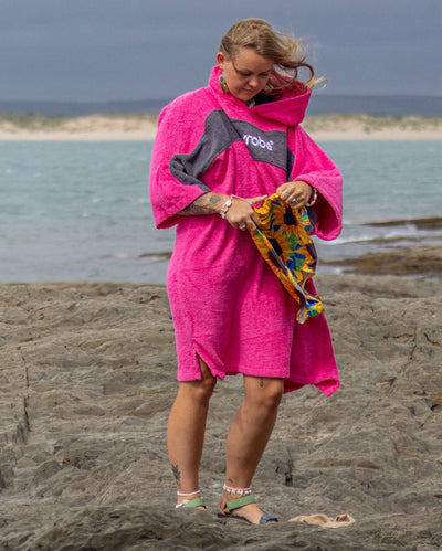 Woman stood by the sea holding a bag, wearing Organic Towel dryrobe Remix in Pink Slate Grey