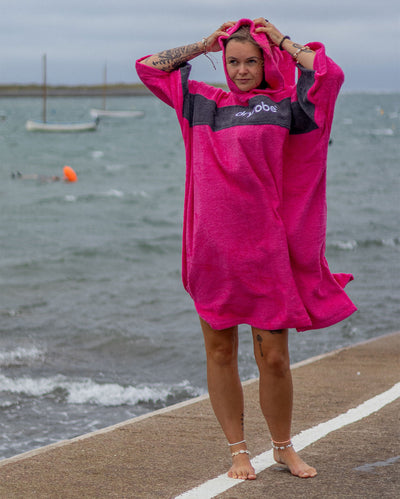 Woman stood on harbour by the sea, wearing Organic Towel dryrobe Remix in Pink Slate Grey with hood up