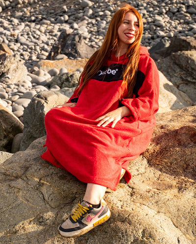 Woman sat on rocks on the beach wearing Organic Towel dryrobe Remix in Red Black