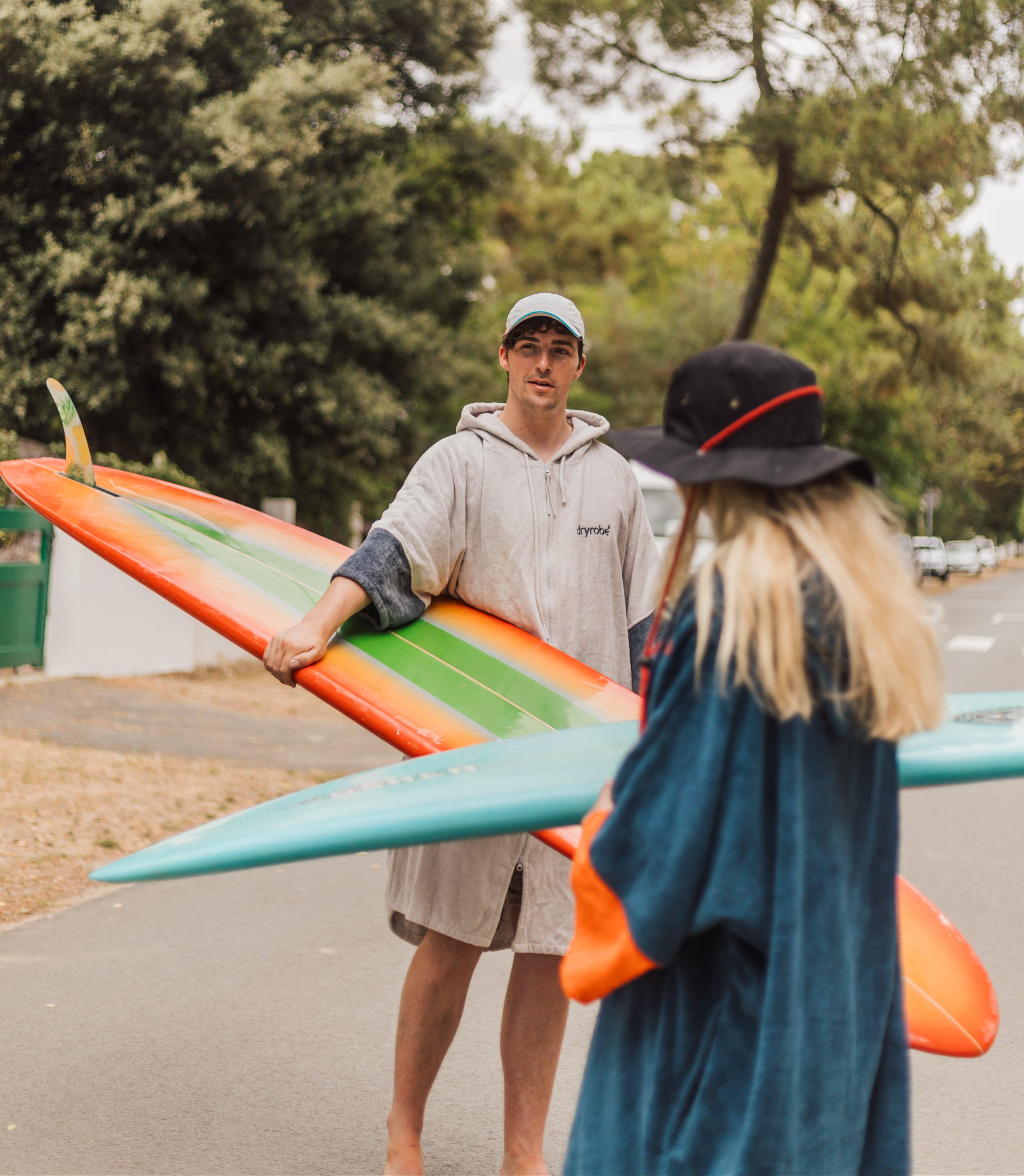 Two people walking with surfboards on a road lined with trees