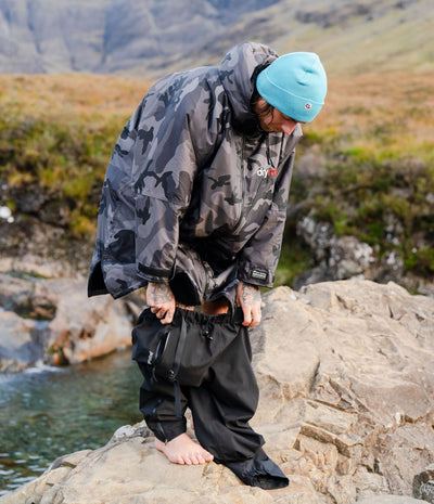*MALE* A person outdoors on a rocky shoreline wearing a dark grey and black camouflage Dryrobe, black pants, and a bright light blue beanie, with mountains and water in the background.