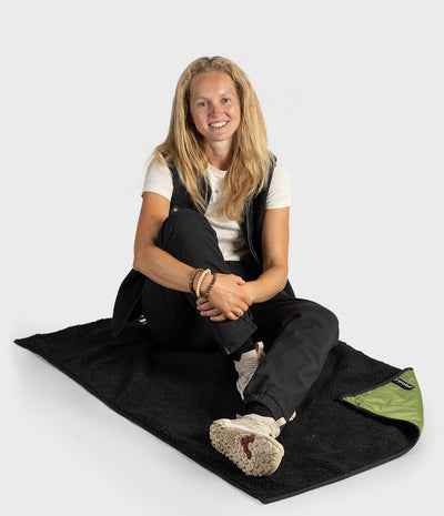 A woman sits cross-legged on a black blanket or sleeping pad with a small green folded corner visible, photographed against a light grey studio background.