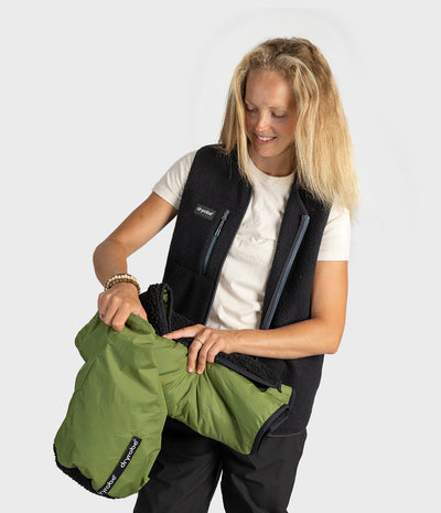 A woman wearing a black fleece vest and white t-shirt holds up an olive green insulated sleeping bag with a black fleece lining, showing the compatibility and fit. The background is a clean, light gray studio backdrop.