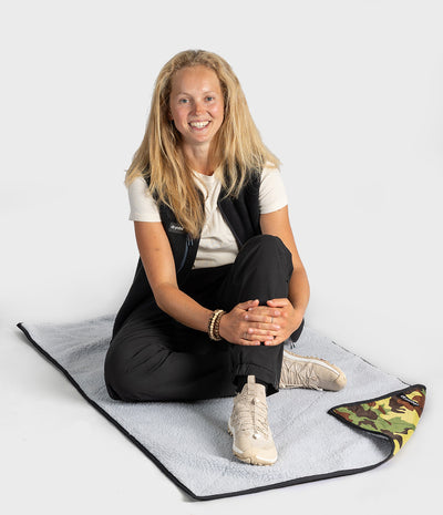 A woman sits cross-legged on a light grey blanket with a black border, showing the reverse side which has a green and yellow camouflage pattern. She is wearing a white t-shirt, a black vest, black pants, and beige sneakers against a plain light grey background.