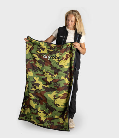 A woman holds up a large camouflage-patterned Dryrobe mat against a light grey studio background. The mat features a green, brown, and yellow camo pattern with the 'dryrobe' logo printed in white near the top left.