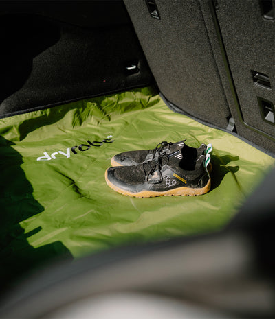 A pair of black and grey trail running shoes with gum soles resting on a bright green Dryrobe floor liner inside a vehicle trunk.