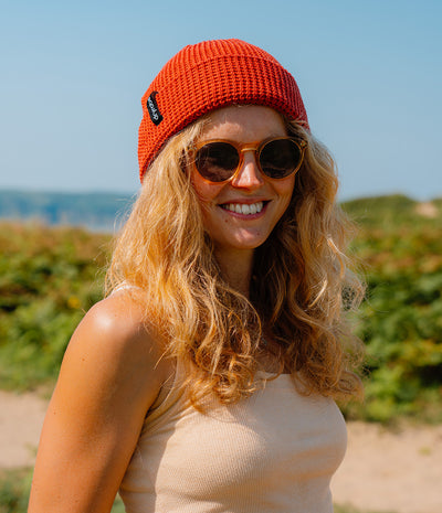 A smiling woman outdoors wearing a bright orange-red knit beanie, round sunglasses, and a light beige ribbed tank top, with blurred greenery and blue sky in the background.