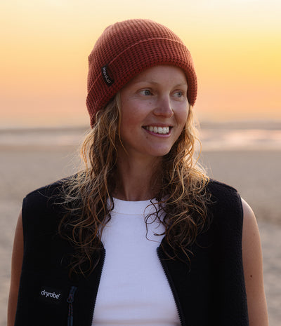 A smiling woman outdoors at sunset wearing a rust-orange knit beanie with a small black logo patch and a black Dryrobe vest over a white top. The background shows a softly blurred beach or shoreline at warm golden hour.