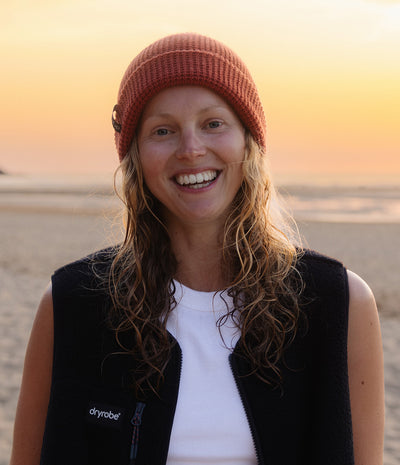 A smiling woman on a beach at sunset wearing a rust-colored knit beanie, a black Dryrobe fleece vest over a white top, with the ocean and warm sky in the background.