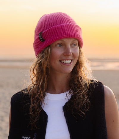 A woman wearing a bright pink knit beanie and a black fleece vest over a white shirt, standing on a beach at sunset.