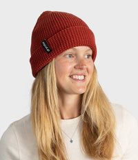 A smiling blonde woman wearing a rust-red ribbed knit beanie with a small black brand tag that reads 'SOLDIER'. The photo is taken against a clean, light gray studio background.