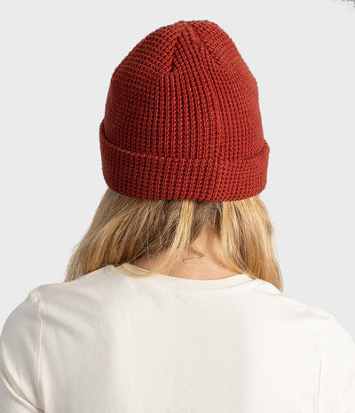 A person viewed from behind wearing a rust-red ribbed knit beanie and a white t-shirt against a light gray background.