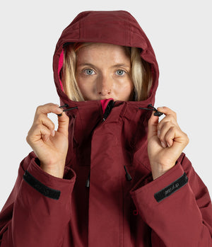 A close-up of a woman wearing a hooded technical jacket in a deep burgundy color, holding the hood up with both hands against a light gray studio background.